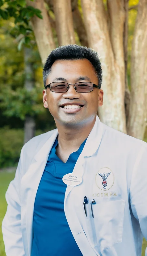 Ceasar Tria, physicians assistant at Coastal Carolina Aesthetics, poses outside smiling in a blue scrubs with a white lab coat over it.