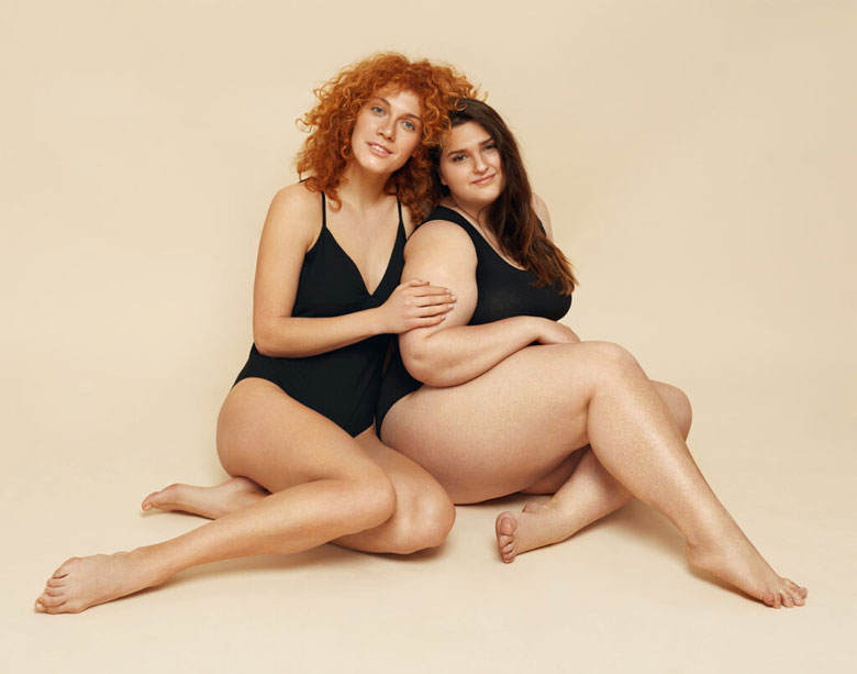 One beautiful woman with curly red hair sits next to a woman with long brown hair. One woman has her hand on the other's arm. Both women have natural bodies and wear black body suits to posing for the benefits of aesthetics treatment at Coastal Carolia.