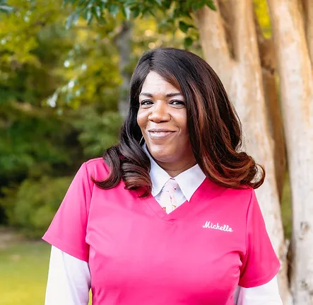 Michelle, a member of the front desk staff at Coastal Carolina Aesthetics, poses outside wearing pink scrubs with a white long-sleeve, collared shirt underneath.