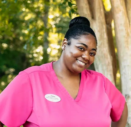 Porsha, Ceasar Tria's medical assistant at Coastal Carolina Aesthetics, poses outside smiling wearing pink scrubs.