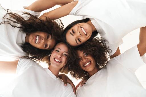 Four women of various ethnicities pose with heads together looking down at the camera with arms around each other's shoulders smile to model the testimonials section of the homepage for Coastal Carolina Aesthetics.